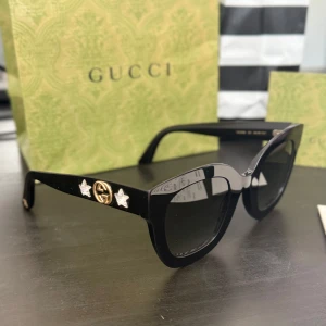 Svarta Gucci solglasögon med stjärndetaljer - Snygga svarta solglasögon från Gucci. På sidorna sitter två glittriga stjärnor och en  GG-logga i guld. Perfekt accessoar för dig som vill addera lite lyx till din stil. Original påse och fodral medföljer samt kvitto och äkthetsbevis. Betalade  350 euro i butiken. PRIS KAN DISKUTERAS!