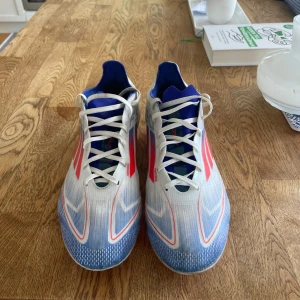 Adiddas F50 fotbollskor - Skriv för funderingar Säljer ett par Adidas fotbollsskor med snygg design i vitt, blått och röda detaljer. Skorna har snörning, spetsig tå och platt sula med dobbar i rosa och vitt. Ovandelen är i syntetmaterial och insidan är fodrad i blått. Perfekta för dig som vill sticka ut på planen!