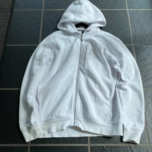 Vit hoodie från Sail Racing, storlek L - Vit hoodie från Sail Racing med dragkedja framtill och broderad logga på bröstet. Tröjan har huva, två sidofickor och en extra bröstficka med dragkedja. Tillverkad i mjuk bomullsmix, perfekt för chill dagar.