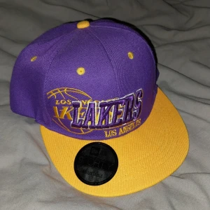 Lakers keps New Era  - Snygg Los Angeles Lakers keps från New Era 9FORTY med broderad logga fram och bak. Kepsen är lila med gula detaljer och gul skärm, justerbar snapback och NBA-märke på sidan. Perfekt för basketfans som vill sticka ut.