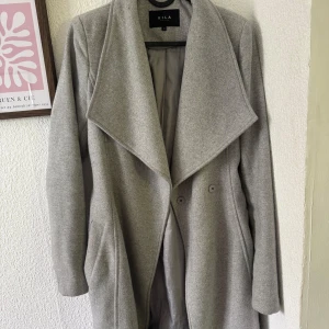 Grå kappa från Vila med bälte XS - Snygg grå kappa från Vila i storlek XS med bred krage och knappar framtill. Kappan har bälte i midjan och är helfodrad. Perfekt för dig som gillar stilrena och klassiska plagg med en modern touch.