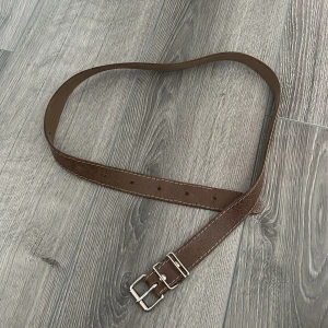 Brunt skärp med dubbla spännen - Snyggt brunt bälte i skinn med dubbla silverspännen och vita kontrastsömmar längs kanterna. Perfekt accessoar för att lyfta din outfit med en klassisk touch. Bältet har flera hål för justerbar passform.