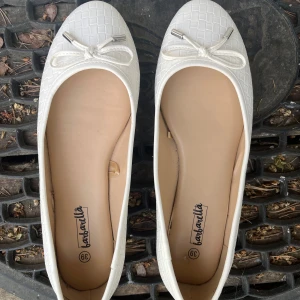 Vita ballerinaskor med rosett från Barbarella - Snygga vita ballerinaskor från Barbarella med flätad struktur och söt rosett framtill med metalländar. Skorna har rund tå och platt sula, perfekta för en clean och fräsch look. Skulle säga att det går mer åt 38/38,5 i storleken