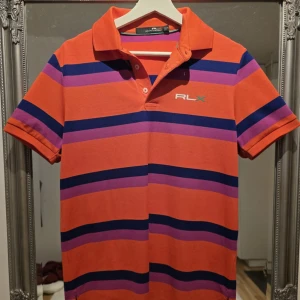 Ralph Lauren - Snygg pikétröja från RLX Ralph Lauren i orange med breda lila och mörkblå ränder. Klassisk krage, kort ärm och broderad RLX-logga på bröstet och ärmen. Tillverkad i bomull för en skön och sportig känsla.