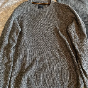 Grå stickad tröja från Lindbergh - Säljer en gråmelerad stickad tröja från Lindbergh. Tröjan har rund halsringning, lång ärm och en klassisk, ribbad struktur. Perfekt för dig som gillar en enkel och stilren look med lite extra textur.