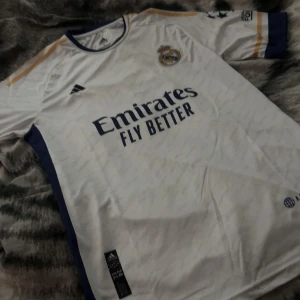 Real Madrid tröja  - Säljer en  Real Madrid fotbollströja från Adidas med Arda Güler och nummer 24 på ryggen. Tröjan är vit med marinblå och guldiga detaljer, korta ärmar och klubbmärke på bröstet. La Liga- och Champions League-märken på ärmarna. Materialet är lätt och andas.