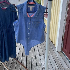 Ärmlös blå skjorta från L.O.G.G. - Snygg ärmlös skjorta från L.O.G.G. i blått med ljusrosa detaljer på insidan av kragen. Skjortan har knappar framtill och en liten bröstficka med rosa sömmar. Perfekt till sommaren och enkel att styla med jeans eller shorts.