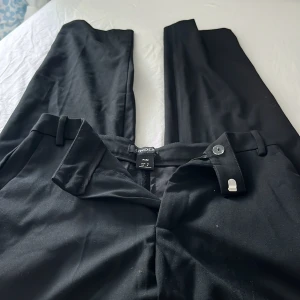 Svarta kostymbyxor Lindex Polly 34 - Snygga svarta kostymbyxor från Lindex, modell Polly. Byxorna har raka ben, hällor för bälte och stängs med knapp och hake. Perfekta för en clean och stilren look. Materialet känns mjukt och följsamt.