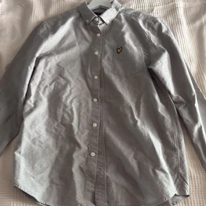 Grå skjorta från Lyle & Scott, storlek M - Snygg grå skjorta från Lyle & Scott med klassisk button-down krage och vita knappar. Bröstficka med gul broderad logga. Skjortan är långärmad och har en stilren, enkel design som passar till många olika looks. Aldrig använd