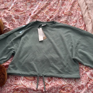 Grön croppad sweatshirt från Noisy May - Snygg ljusgrön croppad sweatshirt från Noisy May med dragsko nertill. Tröjan har långa ärmar och en relaxed fit som ger en chill vibe. Perfekt att matcha med höga jeans eller mjukisbyxor. Materialet är mjuk bomull som känns skönt mot huden.
