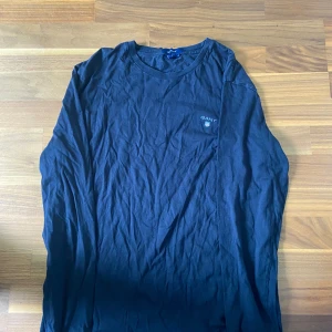 Mörkblå långärmad tröja från GANT - Snygg mörkblå långärmad tröja från GANT med rund halsringning och broderad logga på bröstet. Tillverkad i mjuk bomull som känns skön mot huden. Perfekt för en clean och stilren look.