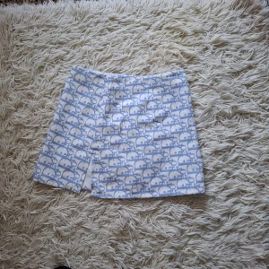 Vit och ljusblå kortkjol med mönster - Superfin kortkjol i vitt och ljusblått med ett unikt, lekfullt mönster. Kjolen har en liten slits framtill och är i en rak modell. Perfekt för dig som vill sticka ut med en cool och söt look.