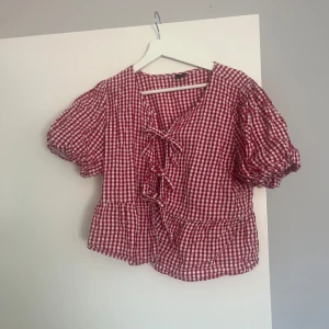 Rutig blus med knyt från Gina Tricot - Superfin röd- och vitrutig blus från Gina Tricot med puffiga korta ärmar och tre söta knyt framtill. Blusen har volangkant nertill. Strl XL