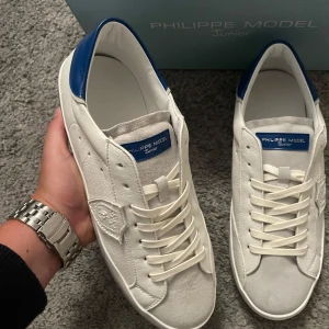 Vita Philippe Model Junior sneakers - Ett par helt nya, vita Philippe Model Junior sneakers med blå detalj på hälen och blå logga på plösen. Skorna har klassisk snörning, rund tå och platt sula. Skick 10/10. (Självklart kan pris diskuteras!)