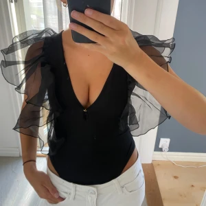 Svart body med volangärmar och djup v-ringning - Supersnygg svart body med transparenta volangärmar i organza och djup v-ringning. Kroppsnära passform som framhäver figuren och ger en cool statement-look. Perfekt för dig som vill sticka ut med en trendig och unik stil.