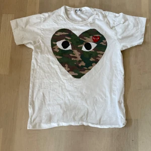 Vit Comme des Garçons Play t-shirt - Vit t-shirt från Comme des Garçons Play med stort hjärta i camouflage-mönster och ögon på bröstet. Liten röd hjärtlogga med ögon på vänster sida. Klassisk rund hals och korta ärmar. Tillverkad i mjuk bomull, perfekt för en cool streetstyle-look.