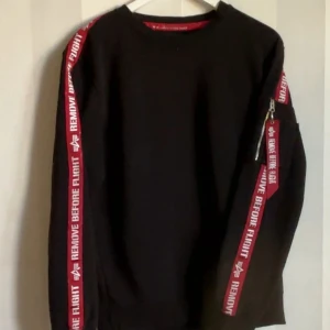 Alpha Industries svart sweatshirt L - Svart sweatshirt från Alpha Industries i storlek L. Snygga röda band med texten 'REMOVE BEFORE FLIGHT' längs ärmarna och dragkedja med detalj på ena ärmen. Klassisk crewneck-modell i mjuk bomull, perfekt för en streetwear-look.