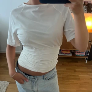 Vit croppad topp med rynkade detaljer - Säljer en vit croppad topp med kort ärm och snygga rynkade detaljer över magen. Toppen har en figurnära passform och är tillverkad i ett mjukt bomullstyg. 
