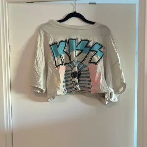 KISS croppad t-shirt H&M L - Cool croppad t-shirt från H&M i storlek L med stort KISS-tryck framtill i blått och svart. T-shirten är vit med lös passform och korta ärmar. Perfekt för dig som gillar bandtröjor och vill sticka ut lite extra.