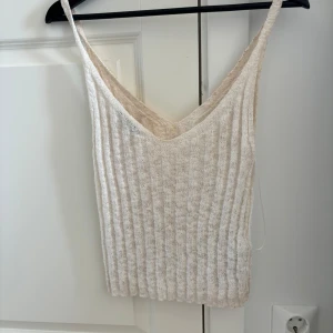 Beige ribbad stickad linnetopp - Supersöt beige stickad linnetopp med ribbad struktur och djup v-ringning. Perfekt för varma dagar och enkel att matcha med jeans eller kjol. Tunn och luftig med smala axelband för en avslappnad vibe.
