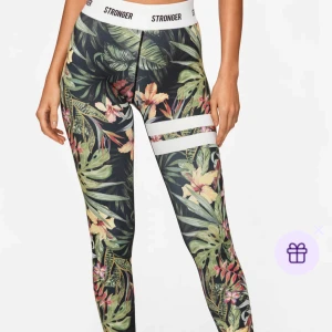 Stronger leggings med tropiskt mönster - Snygga leggings från Stronger i storlek M med tropiskt blommönster i grönt, gult och rosa. De har bred vit resår i midjan med logga och två vita ränder på ena benet. Tillverkade i återvunnen polyester och elastan för en stretchig och bekväm passform. 
