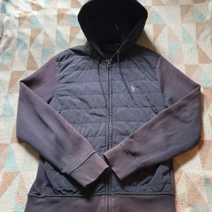 Unik Blå Cardigan från Polo Ralph Lauren - Snygg blå hoodiejacka från Polo Ralph Lauren med quiltad framsida, dragkedja och huva med snörning. Ärmar och rygg i mjuk bomull, framsidan i glansig polyester. Klassisk logga broderad på bröstet. Perfekt för lager-på-lager under höst och vinter. Jackan är väldigt sällsynt och unik. Det som är negativt med jackan är att den är lite solblekt på baksidan men det är inget man direkt märker.