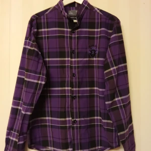Lila rutig skjorta från Ronaldo XL - Snygg lila och svart rutig skjorta från Ronaldo Collection i storlek XL. Skjortan har klassisk krage, långa ärmar och knäppning framtill. Broderad detalj med R på bröstet. Perfekt för dig som gillar färg och mönster. Kan skickas med extra pris via post.