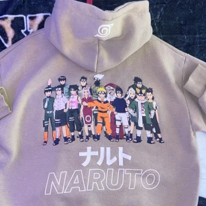 Beige Naruto hoodie med tryck - Beige hoodie med stor Naruto-tryck på ryggen som visar flera karaktärer från serien. Framsidan har Naruto-logga i vitt och ärmen har en vit symbol. Hoodien har huva och känguruficka, perfekt för animefans.