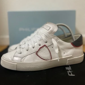 Vita Philippe Model sneakers - Snygga vita sneakers från Philippe Model med klassisk låg siluett. Skorna har detaljer i rött och mörkgrått vid hälen samt en ikonisk sköld på sidan. Tillverkade i skinn med vita snören och platt sula. Perfekta för en clean och trendig look. Mycket bra skick! Storlek 40. Sparsamt använda. Allt org medföljer. Nypris runt 3500!!! Skriv ifall ni har några frågor! 💬 