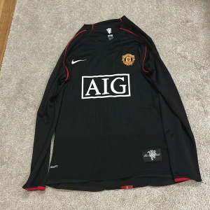 Manchester United långärmad Nike tröja - Snygg svart långärmad Manchester United fotbollströja från Nike med röda detaljer och broderat klubbmärke på bröstet. AIG-logga på framsidan och diskreta ränder i tyget. Tillverkad i lätt och ventilerande material, perfekt för match eller träning.