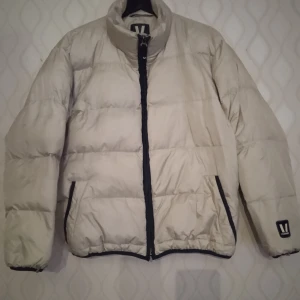 Super snygg beige färg pufferjacka med Vuarnet märke, L - Super snygg beige pufferjacka från Vuarnet med svart dragkedja och detaljer. Jackan har hög krage, två fickor med dragkedja och en klassisk pufferlook. Perfekt för kalla dagar och riktigt skön att bära. Vuarnet-logga på ärmen ger extra stilpoäng. Kan skickas med extra pris via post.