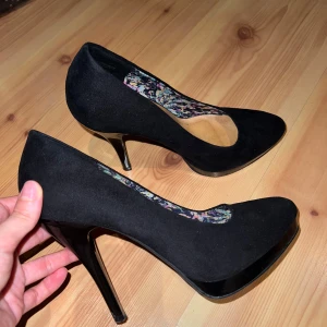 Svarta pumps från DinSko med hög klack - Stilrena svarta pumps från DinSko med hög, smal klack och spetsig tå. Ovandel i mocka-look och insida med färgglatt blommigt mönster. Perfekta för dig som vill ha en klassisk men ändå unik sko med extra höjd.