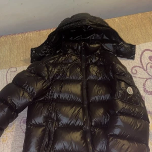 Svart Moncler pufferjacka med huva - Säljer en svart, glansig pufferjacka från Moncler med huva och dragkedja framtill. Jackan har klassisk Moncler-logga på ärmen och stora quiltade partier som ger en riktigt fet vinterlook. Perfekt för dig som vill ha både stil och värme under kalla dagar.
