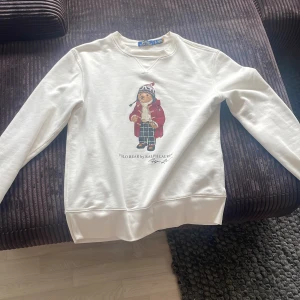 Vit Polo Bear tröja från Ralph Lauren - Vit långärmad sweatshirt från Polo Ralph Lauren med ikoniskt Polo Bear-tryck framtill. Tröjan har rund halsringning, ribbade muddar och är gjord i mjuk bomull. Perfekt för dig som gillar klassisk och lekfull stil. Märks inte att den är använd!