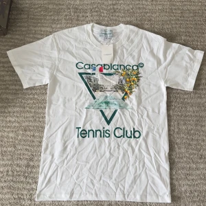 Vit Casablanca Tennis Club t-shirt - Vit t-shirt från Casablanca med stort tryck på bröstet som visar en tennisbana, byggnader och apelsinträd. Grön text med 'Tennis Club' och cool grafisk design. Klassisk passform och rund hals. Perfekt för dig som gillar sportig och trendig stil.