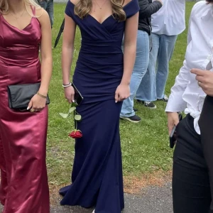 Marinblå långklänning offshoulder - Säljer en marinblå långklänning med offshoulder och v-ringad omlott över bysten. Klänningen har figurnära passform och är sydd i ett slätt, stretchigt material som ger en elegant siluett. Perfekt för bal eller student! 1000kr nypris använd en gång