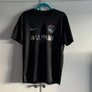Svart PSG x Balmain fotbollströja Nike M - Tjena säljer nu en snygg svart PSG fotbollströja i Dri-FIT-material från Nike, med holografiskt Balmain Paris-tryck och PSG-logga på bröstet. Klassisk rund hals och korta ärmar. Perfekt för dig som vill sticka ut på planen eller gatan. OBS tröjan är ALDRIG använd!