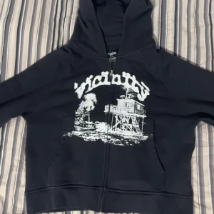 hoodie från Vicinity shooting house - Svart hoodie från Vicinity med stor vit grafisk print framtill. Tröjan har huva, dragkedja. Perfekt för dig som gillar streetwear vibes och vill sticka ut med en unik design. Helt ny med tag aldrig använd 