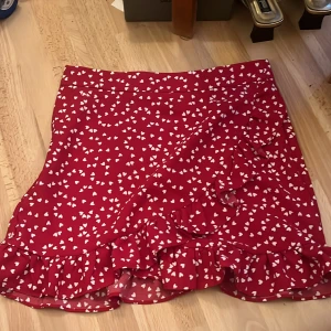 Röd omlottkjol med volang och hjärtan - Supersöt röd kortkjol med vita hjärtan, omlottmodell och volangkant. Perfekt för dig som vill sticka ut med en lekfull och trendig look. Kjolen har en lätt och luftig känsla, perfekt för varmare dagar.