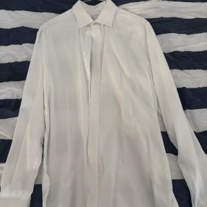 Vit skjorta från Bläck, tailored fit - Klassisk vit skjorta från Bläck i tailored fit. Skjortan har lång ärm, knappar framtill och en stilren krage. Perfekt för dig som gillar en clean och tidlös look. Materialet är bomull som känns skönt mot huden.