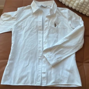 Vit skjorta från U.S. Polo Assn, stl 36 - Klassisk vit skjorta från U.S. Polo Assn. med broderad logga på bröstet. Skjortan har lång ärm, knappar framtill och är tillverkad i en skön mix av bomull och polyester. Perfekt för en clean och stilren look.