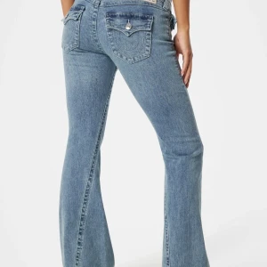 True Religion Joey Low Rise Flare Jeans - Säljer ett par ljusblå True Religion Joey jeans med låg midja och utsvängda ben. Klassiska bakfickor med lock och silverfärgade knappar med logga. Jeansen är i bomull och har tydliga kontrastsömmar för en snygg vintagekänsla.