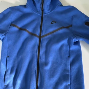 Blå Nike tech fleece  - Snygg blå techfleece från Nike med svart logga på låret och bröstet. Byxorna och tröjan har en sportig look, riktigt sköna att ha på sig när man bara tar de lungt eller om man ska träna. 