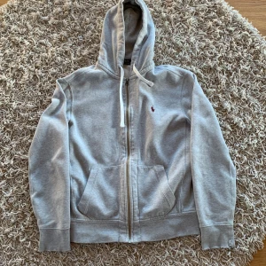 Grå hoodie från Polo Ralph Lauren M - Klassisk grå hoodie från Polo Ralph Lauren i storlek M. Tröjan har dragkedja, huva med snörning och känguruficka framtill. Liten broderad logga på bröstet. Perfekt för chill dagar och enkel att matcha med jeans eller joggers. Titta bild 4&5.