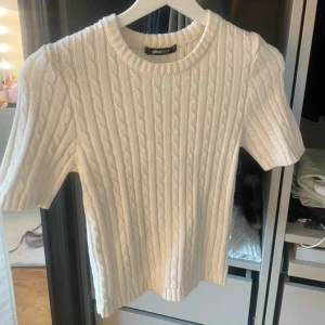 Vit kabelstickad tröja Gina Tricot - Vit kabelstickad tröja från Gina Tricot med korta ärmar och rund halsringning. Tröjan har en klassisk kabelstickad struktur och är gjord i ett mjukt material som känns skönt mot huden. Perfekt för lager-på-lager och snygg till både jeans och kjol.