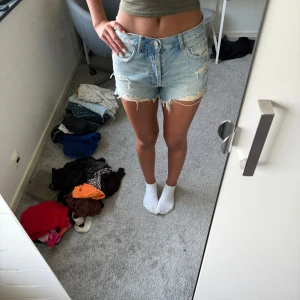 Ljusblå jeansshorts med slitningar - Säljer ett par ljusblå jeansshorts med råa kanter och slitna detaljer. Klassisk femficksmodell med knapp och dragkedja framtill. Shortsen har en midwaist passform och är perfekta för varma dagar. Snyggt avslappnad stil som passar till allt.