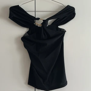 Svart offshoulder topp med knutdetalj - Trendig svart offshoulder topp med cool knutdetalj framtill. Toppen är tight och har en unik skärning över axlarna som ger en snygg siluett. Perfekt för dig som vill sticka ut med en stilren men edgy look.