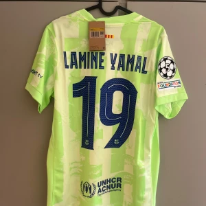 Barcelona Lamine Yamal matchtröja Nike - Säljer en Barcelona fotbollströja med Lamine Yamal 19 på ryggen. Tröjan är ljusgrön med vita detaljer, har korta ärmar och är tillverkad i lätt, ventilerande material. Tryck från Spotify, UNHCR och UEFA Foundation samt klubbmärke och Nike-logga på bröstet.