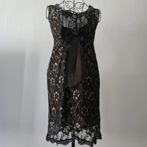 Svart spetsklänning med guld och rosett - Supersnygg svart kortklänning med guldiga detaljer och spets över hela klänningen. Framtill sitter en stor svart rosett som ger extra wow-känsla. Klänningen är ärmlös och har en figurnära passform.