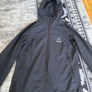 Svart vindjacka från Haglöfs med huva - Säljer en svart vindjacka från Haglöfs med snygg minimalistisk design. Jackan har hel dragkedja framtill, två dragkedjeförsedda fickor och en praktisk huva. Perfekt för blåsiga dagar och outdoor-äventyr. Lätt och smidig att bära med sig.